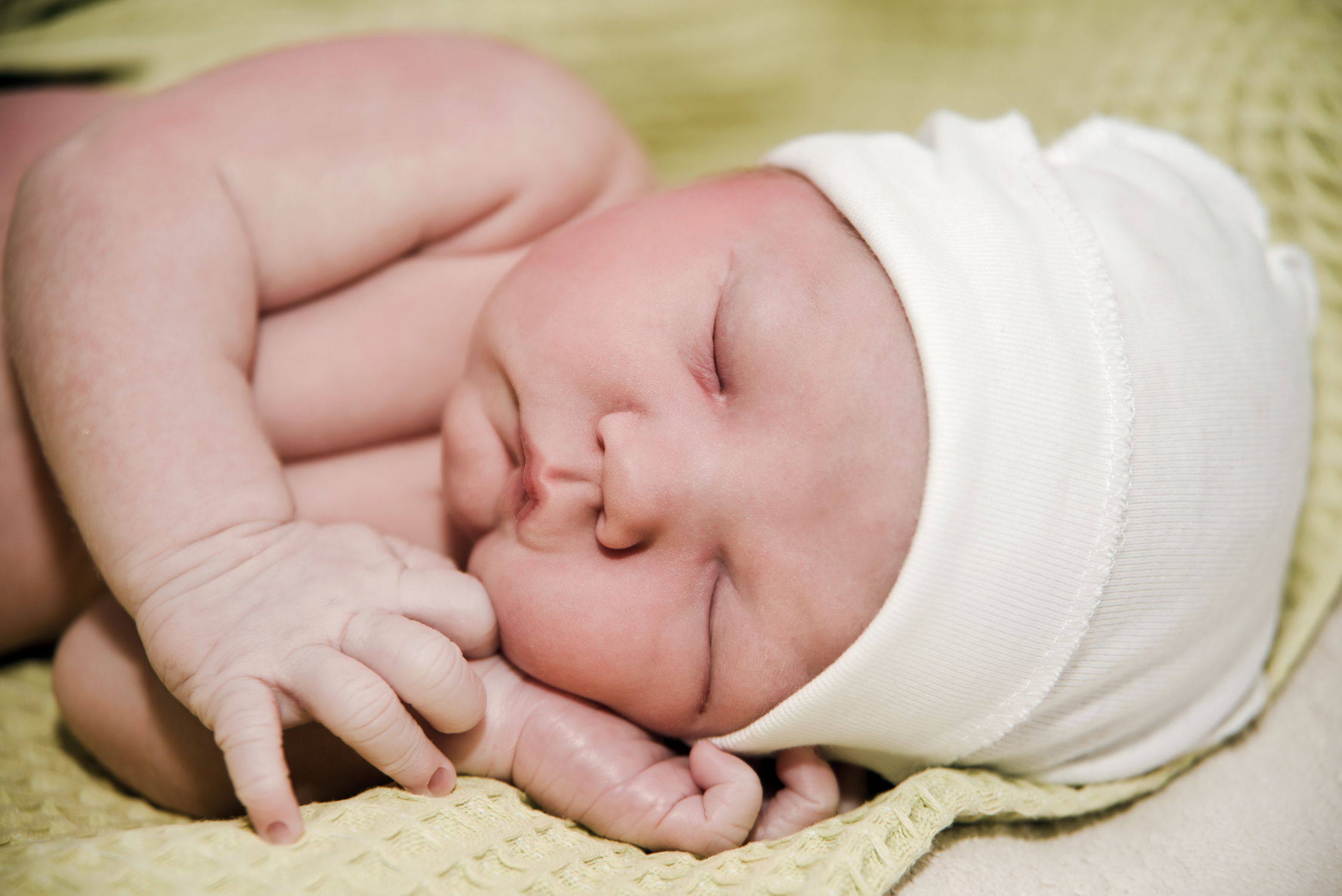 Sleeping newborn in a white hat, laying on their left side on a yellow green blanket